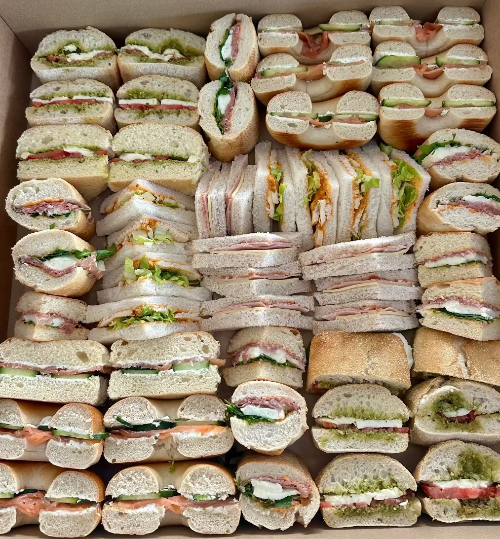 Box filled with various types of sandwiches including bagels and ciabatta. Ham and cheese, cajun chicken, pesto and mozerella, salami and rocket. 